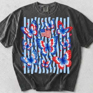 Puede incluir: Una camiseta de manga corta gris carb&oacute;n con un estampado gr&aacute;fico de varias mariposas con patrones de alas en rojo, blanco y azul. El dise&ntilde;o incluye una peque&ntilde;a bandera estadounidense y rayas verticales azul claro con estrellas rojas y azules.