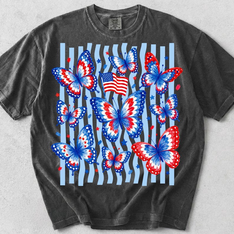 Puede incluir: Una camiseta de manga corta gris carb&oacute;n con un estampado gr&aacute;fico de varias mariposas con patrones de alas en rojo, blanco y azul. El dise&ntilde;o incluye una peque&ntilde;a bandera estadounidense y rayas verticales azul claro con estrellas rojas y azules.