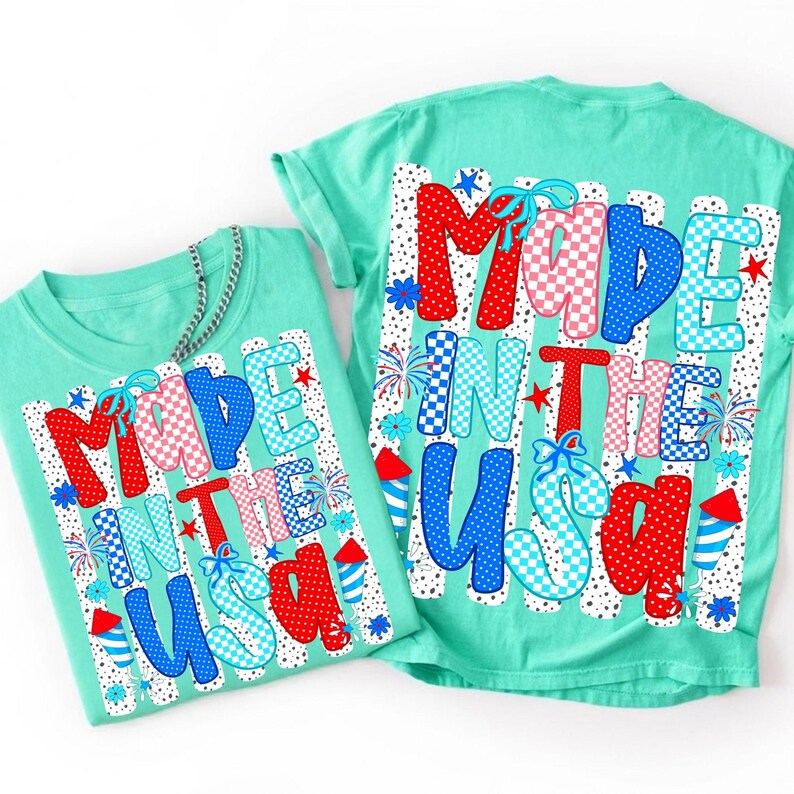May include: A mint green t-shirt featuring a graphic print with the text MADE IN THE USA. The letters are filled with red, white, and blue patterns including polka dots and checkers. The design includes small firework and rocket illustrations.