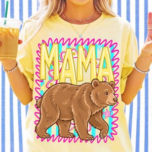 May include: A pale yellow t-shirt featuring a graphic print of a brown bear walking in front of the word MAMA in large yellow letters. The design is framed by a bright pink squiggly border and includes small blue and pink star accents.