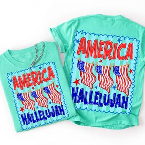 Puede incluir: Una camiseta verde menta con un estampado gr&aacute;fico de tres banderas estadounidenses colgadas en un tendedero. El texto AMERICA aparece en rojo sobre las banderas y HALLELUJAH en azul debajo, todo enmarcado por un borde azul festoneado.