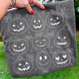 May include: A dark grey fabric tote bag featuring a white line art pattern of various jack-o'-lanterns with different facial expressions, including smiling, heart-shaped eyes, and jagged teeth, interspersed with small decorative bows and stars.