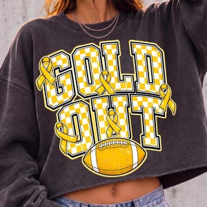 Puede incluir: Una sudadera corta de color gris carb&oacute;n con el texto GOLD OUT en un patr&oacute;n de cuadros amarillos y blancos. El dise&ntilde;o incluye cuatro lazos amarillos de concienciaci&oacute;n y un gr&aacute;fico de un bal&oacute;n de f&uacute;tbol americano amarillo con rayas blancas.