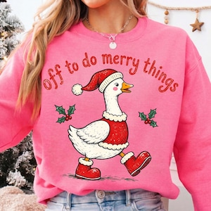 May include: A bright pink crewneck sweatshirt featuring a graphic of a white goose wearing a red Santa hat, a red vest with white trim, and red boots. The text "off to do merry things" is printed in red above the goose, with small holly sprigs.