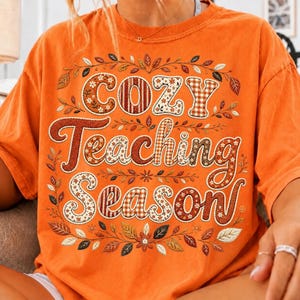 May include: An orange short-sleeve t-shirt featuring the text "Cozy Teaching Season" in a large, decorative font. The letters are filled with various patterns including plaid, floral, and solid colors, surrounded by a border of autumn leaves.