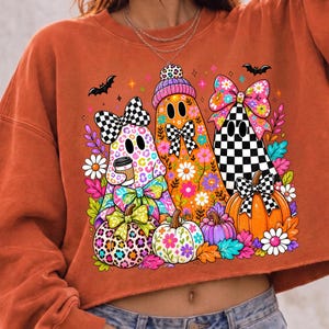 Puede incluir: Una sudadera de color naranja quemado con un gr&aacute;fico de tres fantasmas decorados con patrones coloridos, incluyendo estampado de leopardo, dise&ntilde;os florales y un tablero de ajedrez blanco y negro. Los fantasmas est&aacute;n rodeados de calabazas, murci&eacute;lagos y flores.