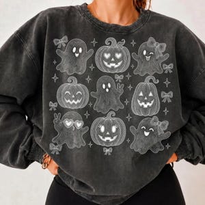 Puede incluir: Una sudadera de cuello redondo en color gris carb&oacute;n con un estampado gr&aacute;fico de l&iacute;neas blancas. El dise&ntilde;o muestra una cuadr&iacute;cula de tres calabazas y seis fantasmas, decorada con peque&ntilde;as estrellas y lazos. Los fantasmas y calabazas tienen diversas expresiones faciales.