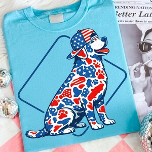 Puede incluir: Una camiseta azul claro con el gr&aacute;fico de un perro sentado que lleva una gorra de b&eacute;isbol. El perro tiene un estampado de estrellas y huellas de patas en rojo, blanco y azul. Un contorno azul oscuro en forma de rombo rodea la ilustraci&oacute;n.