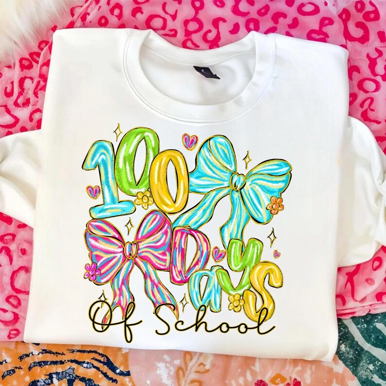 Op de afbeelding: Witte sweatshirt met een kleurrijke afbeelding ter viering van "100 Days Of School". Het ontwerp toont de cijfers "100" in groen en geel, met de woorden "Days" en "Of School" in een speels lettertype. Kleurrijke strikken en harten dragen bij aan het feestelijke thema.