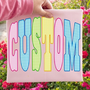 May include: A light pink fabric square featuring the word CUSTOM printed in large, bold, block letters. Each letter displays a vertical striped pattern in alternating colors of pink, blue, green, and yellow with a dark blue outline.