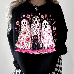 May include: A black sweatshirt featuring a graphic print of three white ghosts decorated with pink flowers, leopard spots, and polka dots. The design includes black cats and pink awareness ribbons scattered throughout the illustration.