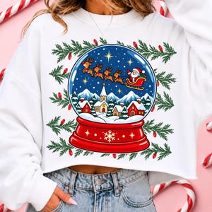 May include: A white long-sleeve sweatshirt featuring a graphic print of a snow globe containing a winter village scene with a church, houses, and Santa Claus in a sleigh pulled by reindeer. The design is framed by green pine branches and red lights.