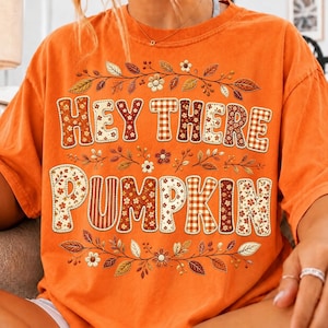 May include: An orange short-sleeve t-shirt featuring the text "HEY THERE PUMPKIN" in large, blocky letters filled with various brown, orange, and white floral and plaid patterns, surrounded by decorative illustrations of leaves and small flowers.