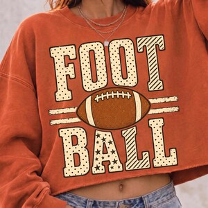 May include: A burnt orange cropped sweatshirt featuring a large graphic print with the word FOOTBALL in cream-colored block letters. The letters are filled with polka dots, stars, and diagonal stripes. A brown football is centered between the words.