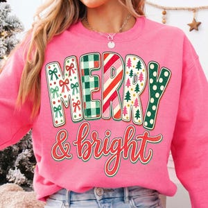 May include: A bright pink crewneck sweatshirt featuring the text MERRY in large block letters with varied holiday patterns including red bows, green plaid, candy cane stripes, small pine trees, and green polka dots, with & bright in red script.