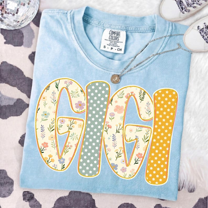 May include: A light blue crew neck t-shirt with a size small Comfort Colors label at the collar. The front features the word GIGI in large block letters. The letters G, G, and I have a floral pattern, while the I has a star pattern.