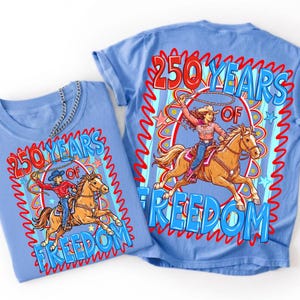 Puede incluir: Una camiseta azul claro con un estampado gr&aacute;fico de una persona montando un caballo marr&oacute;n mientras hace girar un lazo. El texto 250 YEARS OF FREEDOM est&aacute; impreso en rojo y azul. El dise&ntilde;o incluye un borde ondulado rojo y estrellas rosas.