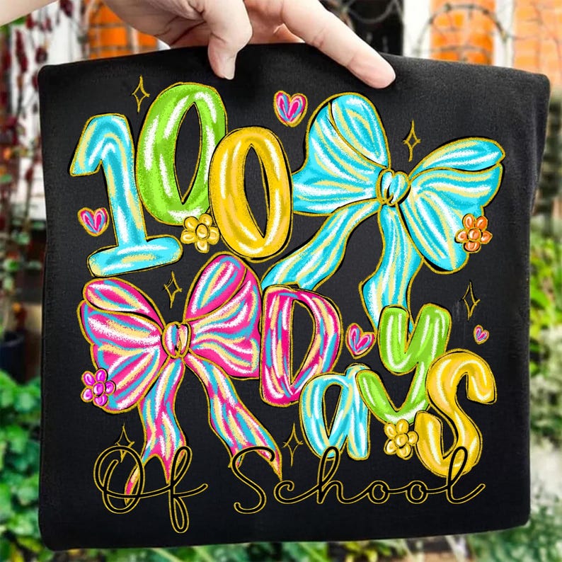 Op de afbeelding: Zwart T-shirt met een kleurrijk ontwerp dat "100 Dagen School" viert. Het ontwerp toont grote cijfers, strikken, harten en bloemen in tinten blauw, groen, geel en roze. De tekst is goudkleurig omlijnd.