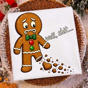 May include: A white t-shirt featuring a cartoon gingerbread person with a worried expression, a green bowtie, and red and green buttons. The gingerbread person has a broken leg with crumbs scattered below and the text well, shit... printed.