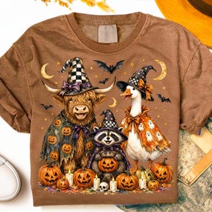 May include: A brown t-shirt featuring a graphic of a highland cow, a raccoon, and a goose wearing witch hats and Halloween costumes. The design includes pumpkins, ghosts, bats, and crescent moons scattered around the animals on the shirt.