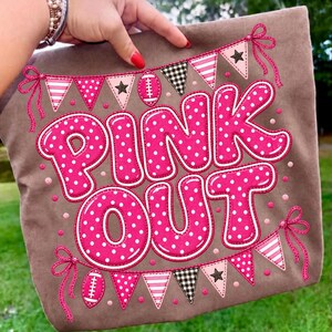 K&ouml;nnte beinhalten: Ein brauner Stoffartikel mit dem Text PINK OUT in gro&szlig;en, leuchtend pinken Buchstaben mit wei&szlig;en Punkten. Das Design enth&auml;lt dekorative Wimpel in Pink, Wei&szlig; und Schwarz sowie kleine Football-Symbole und Schleifen.