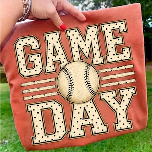 May include: A burnt orange t-shirt featuring the text GAME DAY in large, block-style letters with a black polka dot pattern. A realistic baseball graphic with visible red stitching is positioned in the centre, replacing the letter A.