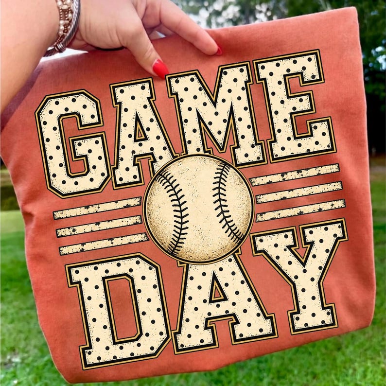 May include: A burnt orange t-shirt featuring the text GAME DAY in large, block-style letters with a black polka dot pattern. A realistic baseball graphic with visible red stitching is positioned in the centre, replacing the letter A.