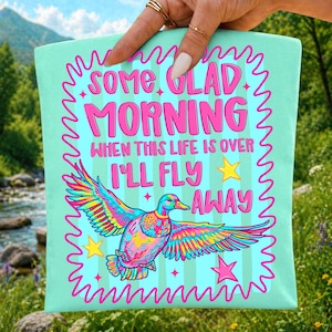 Pu&ograve; includere: Una borsa tote color verde menta chiaro con un'anatra multicolore in volo. Il testo recita "SOME GLAD MORNING WHEN THIS LIFE IS OVER I'LL FLY AWAY", circondato da un bordo rosa ondulato e da stelle rosa e gialle come decorazioni.