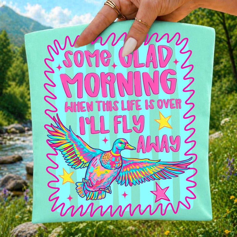 Pu&ograve; includere: Una borsa tote color verde menta chiaro con un'anatra multicolore in volo. Il testo recita "SOME GLAD MORNING WHEN THIS LIFE IS OVER I'LL FLY AWAY", circondato da un bordo rosa ondulato e da stelle rosa e gialle come decorazioni.