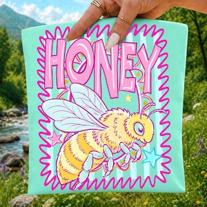 May include: A light mint green tote bag featuring a large, cartoon-style yellow and black striped bee in the center. Above the bee, the word HONEY is printed in bold, bubble-style pink letters. The design is framed by a jagged, bright pink border.