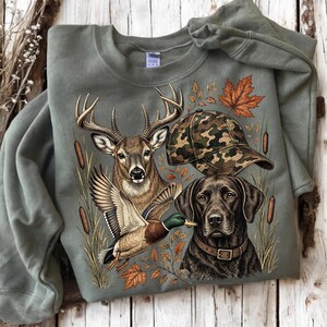 May include: An olive green crewneck sweatshirt featuring a graphic print of a deer, a mallard duck, a black Labrador retriever wearing a collar, and a camouflage baseball cap, all surrounded by cattails and orange autumn leaves.