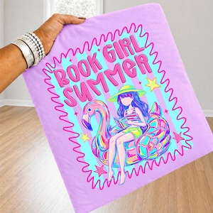 May include: A light purple fabric item featuring a graphic print of a person with long hair wearing a yellow hat, sitting on a pink flamingo pool float while reading a book. The text BOOK GIRL SUMMER is printed in pink above the illustration.