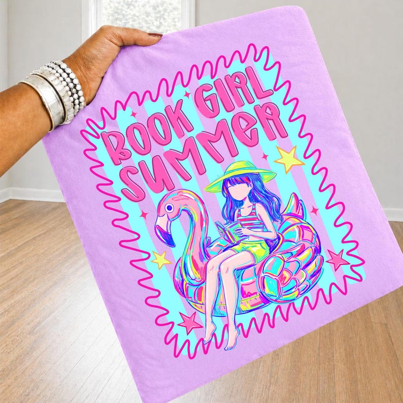 May include: A light purple fabric item featuring a graphic print of a person with long hair wearing a yellow hat, sitting on a pink flamingo pool float while reading a book. The text BOOK GIRL SUMMER is printed in pink above the illustration.