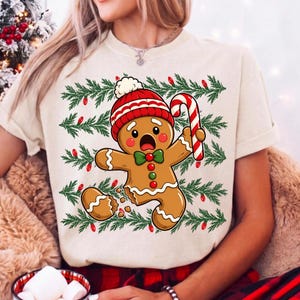 May include: A cream-coloured t-shirt featuring a graphic of a surprised gingerbread person wearing a red and white knit hat and a green bow tie. The gingerbread person holds a red and white candy cane, set against a background of green pine branches with small red Christmas lights.