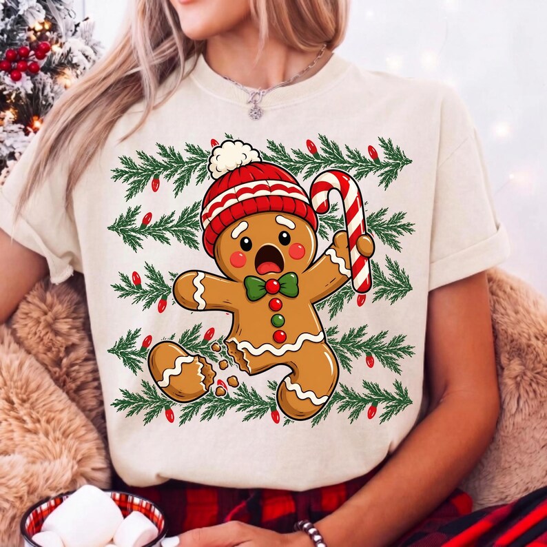 May include: A cream-coloured t-shirt featuring a graphic of a surprised gingerbread person wearing a red and white knit hat and a green bow tie. The gingerbread person holds a red and white candy cane, set against a background of green pine branches with small red Christmas lights.