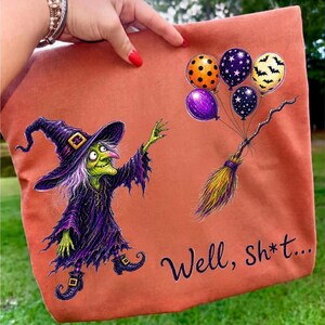 K&ouml;nnte beinhalten: Eine orangefarbene Tragetasche mit einer Grafik einer gr&uuml;nh&auml;utigen Hexe in violettem Gewand und Hut, die nach einem Besenstiel greift, an dem vier Luftballons mit orangefarbenen, violetten und schwarzen Mustern befestigt sind. Der Text "Well, sh*t..." ist schwarz gedruckt.