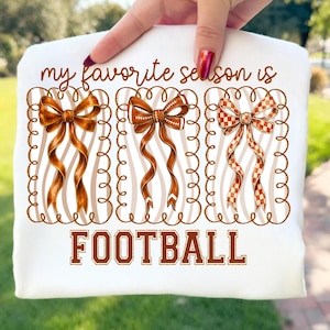 Può includere: Felpa bianca con la scritta "my favorite season is FOOTBALL". Il design presenta tre fiocchi marroni con motivi diversi, su sfondo a righe. I fiocchi sono incorniciati da un bordo decorativo.