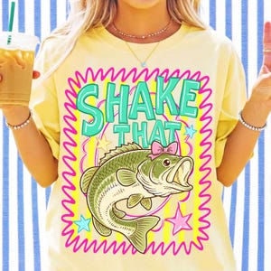 K&ouml;nnte beinhalten: Ein hellgelbes T-Shirt mit einem Grafikdruck, der den Text SHAKE THAT in t&uuml;rkisfarbenen Buchstaben zeigt. Unter dem Text befindet sich ein gr&uuml;ner Fisch mit einer kleinen rosa Schleife. Das Design ist von einem pinken Wellenrand umgeben.