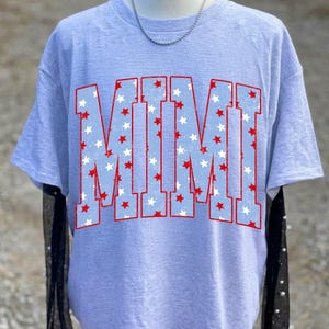 May include: A light gray short-sleeve t-shirt featuring the word MIMI in large block letters. The letters have a light blue background filled with a pattern of small red and white stars, all outlined in a thin red border.
