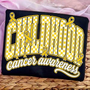 Puede incluir: Una camiseta negra con la palabra CHILDHOOD en letras grandes de bloque a cuadros amarillos y blancos. El texto est&aacute; decorado con varios lazos amarillos. Debajo, las palabras cancer awareness est&aacute;n escritas en una fuente cursiva blanca.