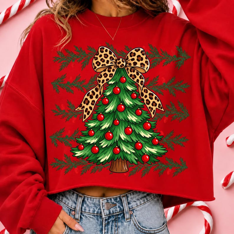Op de afbeelding: Een rood cropped sweatshirt met een afbeelding van een groene kerstboom versierd met rode kerstballen en bekroond met een grote strik met luipaardprint. De achtergrond van de afbeelding bevat horizontale rijen groene dennentakken.