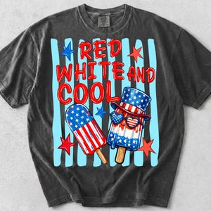 May include: A charcoal grey short-sleeve t-shirt featuring a graphic of two popsicles with red, white, and blue star and stripe patterns. The text reads RED WHITE AND COOL in red letters. The background has light blue vertical stripes and red stars.