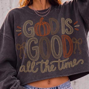 May include: A charcoal gray cropped sweatshirt featuring a graphic design with the text "GOD IS GOOD all the time" in a retro, multi-line font. The design includes a central orange pumpkin illustration and two decorative line-art bows on the sides.