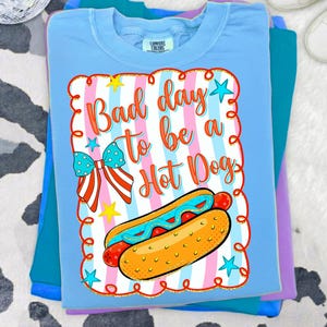 May include: A light blue t-shirt featuring a graphic print with the text "Bad day to be a Hot Dog" in orange script. The design includes a hot dog, a red, white, and blue patterned bow, and yellow stars set against a background of vertical stripes.