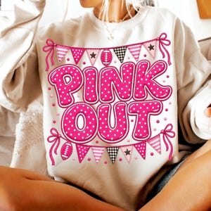 K&ouml;nnte beinhalten: Ein cremefarbenes Sweatshirt mit Rundhalsausschnitt und einem Grafikdruck mit dem Text "PINK OUT" in gro&szlig;en, pinkfarbenen Buchstaben mit wei&szlig;en Punkten. Das Design enth&auml;lt pinkfarbene und schwarz-wei&szlig; gemusterte Wimpel, Sterne und Football-Symbole.