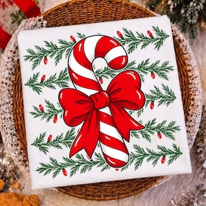 May include: A white square textile featuring a central red and white striped candy cane tied with a large red bow. The background is decorated with horizontal rows of green pine branches interspersed with small red Christmas light bulbs.