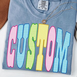 May include: A blue t-shirt with a crew neckline and a Comfort Colors label at the collar. The front features the word CUSTOM in large, multi-colored letters with vertical white stripes in pink, blue, green, yellow, and pink. A thin silver necklace is visible.