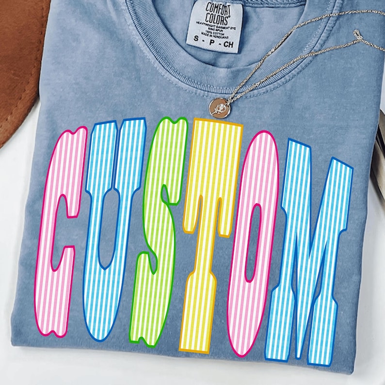 May include: A blue t-shirt with a crew neckline and a Comfort Colors label at the collar. The front features the word CUSTOM in large, multi-colored letters with vertical white stripes in pink, blue, green, yellow, and pink. A thin silver necklace is visible.