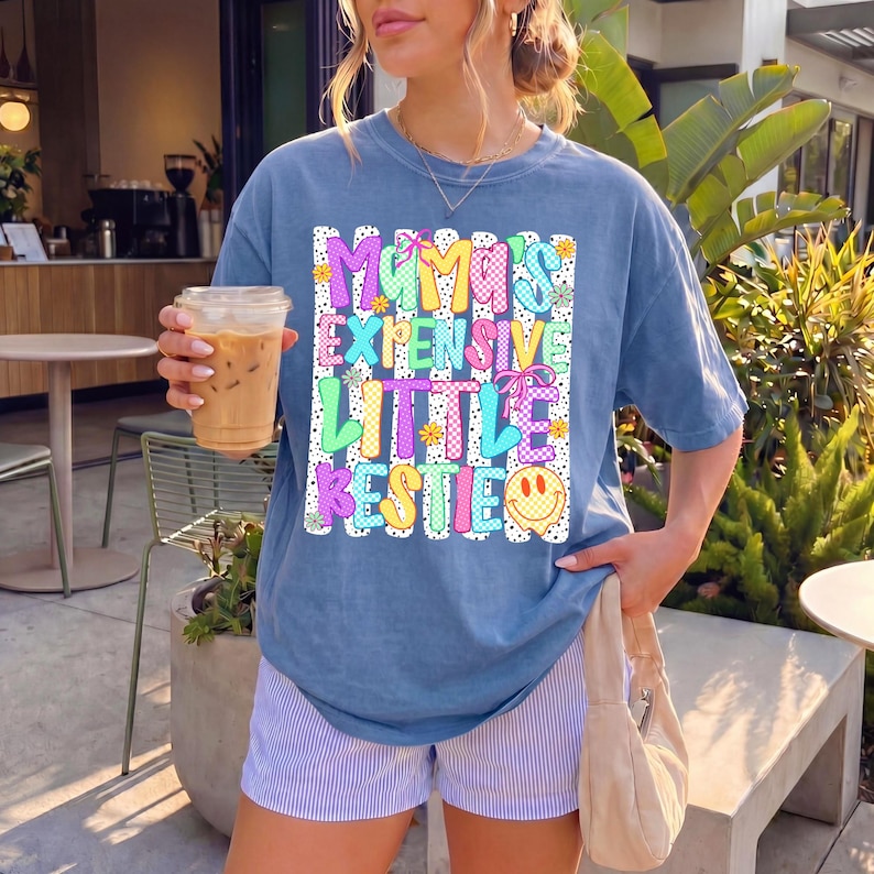May include: A blue short-sleeve t-shirt featuring a graphic print with the text "MAMA'S EXPENSIVE LITTLE BESTIE" in colorful, patterned block letters. The design includes small flowers, bows, and a yellow smiley face on a white striped background.