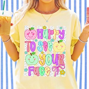K&ouml;nnte beinhalten: Ein blassgelbes T-Shirt mit einem Grafikdruck, der den Text "Happy TO SEE YOUR FACE" in bunten, gemusterten Buchstaben zeigt. Das Design enth&auml;lt drei l&auml;chelnde Apfelillustrationen mit Karomuster sowie kleine blaue und orangefarbene Blumen.